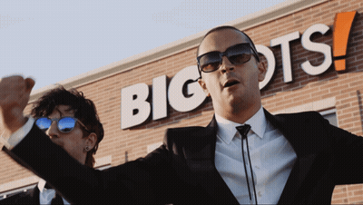 A Man In A Suit Stands In Front Of A Big Ots Sign GIF