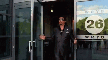 A Man In A Suit Stands In Front Of A Building That Says Wryo 26 GIF