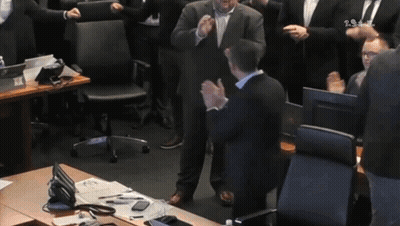 A Man In A Suit Stands In Front Of A Group Of People Applauding With The Letters Ts On The Bottom Left GIF