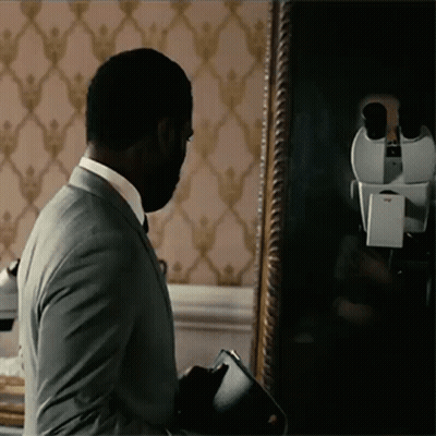 A Man In A Suit Stands In Front Of A Mirror Looking At His Reflection GIF
