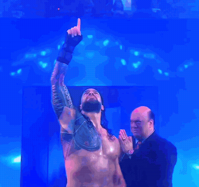 A Man In A Suit Stands Next To A Shirtless Wrestler Who Is Pointing Up At The Sky GIF