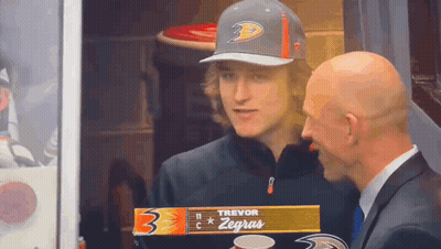 A Man In A Suit Talks To A Young Man Wearing A Hat That Says Trevor Zegras GIF