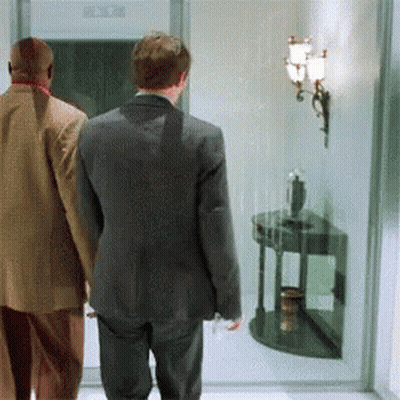 A Man In A Suit Walks With Another Man In A Tan Jacket GIF