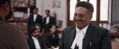 A Man In A Suit With The Letter M On His Lapel Smiles GIF