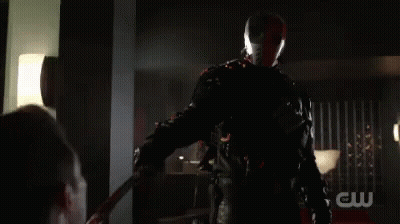 A Man In A Superhero Costume Is Holding A Sword In His Hand In A Dark Room . GIF