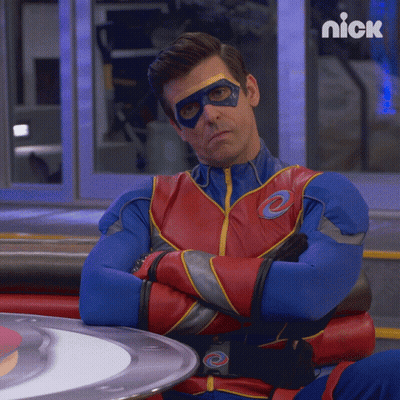 A Man In A Superhero Costume Is Sitting At A Table With His Arms Crossed And A Nick Logo Behind Him GIF