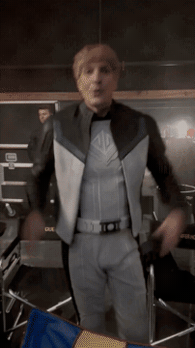 A Man In A Superhero Costume Is Standing In Front Of A Chair With A Blue And Yellow Flag GIF