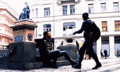 A Man In A Superhero Costume Is Talking To A Man Sitting On The Ground GIF