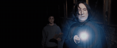 A Man In A Sweater And Tie Stands In A Dark Room With A Light Shining On Him GIF