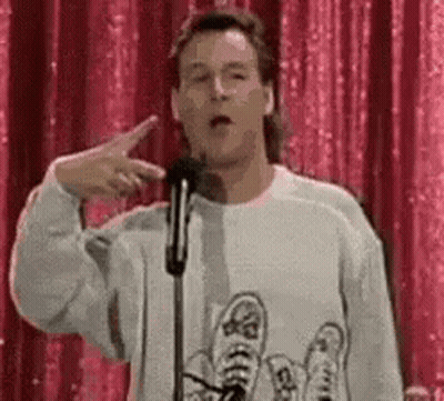 A Man In A Sweater Is Giving A Speech In Front Of A Microphone And Pointing At The Camera . GIF