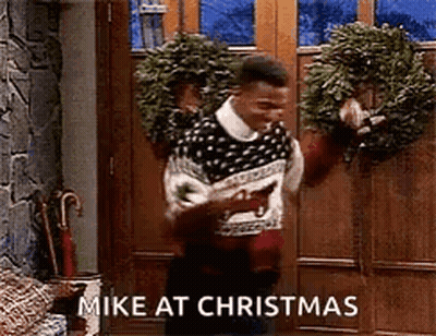A Man In A Sweater Is Standing In Front Of A Door Holding A Wreath And Says `` Mike At Christmas '' GIF