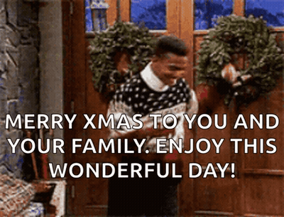 A Man In A Sweater Is Standing In Front Of A Door With A Wreath On It And Says Merry Xmas GIF