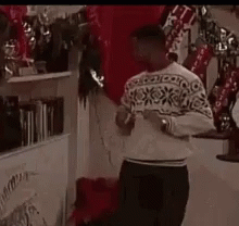 A Man In A Sweater Is Standing In Front Of A Fireplace In A Room . GIF