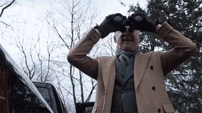 A Man In A Tan Coat Looks Through Binoculars In Front Of A Car GIF