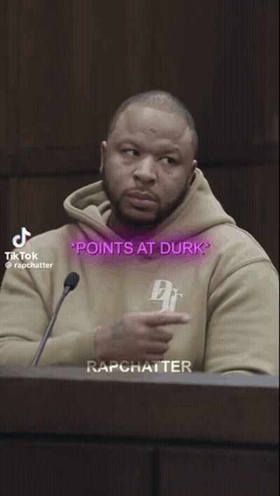 A Man In A Tan Hoodie Points At The Words Points At Durk On His Shirt GIF