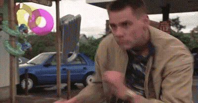 A Man In A Tan Jacket And Tie Is Sitting In Front Of A Blue Car And Inflatable Donuts . GIF