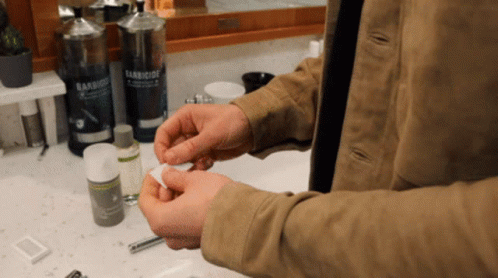 A Man In A Tan Jacket Is Holding A Cotton Ball In Front Of A Bathroom Sink With A Bottle Of Baniocide On The Counter GIF