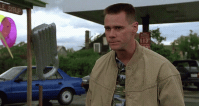 A Man In A Tan Jacket Stands In Front Of A Gas Station With A Sign That Says 113 GIF