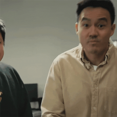 A Man In A Tan Shirt Is Making A Funny Face While Standing Next To Another Man In A Green Sweater . GIF