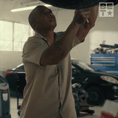A Man In A Tan Shirt Is Working On A Tire In A Garage With The Word Be On The Bottom Right GIF