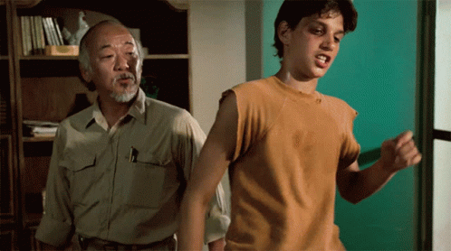 A Man In A Tan Shirt Stands Next To Another Man In A Yellow Shirt GIF