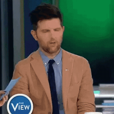 A Man In A Tan Suit And Tie Is Holding A Piece Of Paper In Front Of A Sign That Says The View . GIF