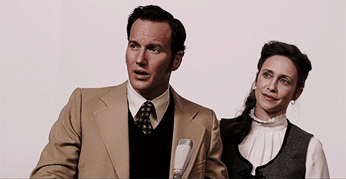 A Man In A Tan Suit And Tie Stands Next To A Woman In A White Shirt GIF