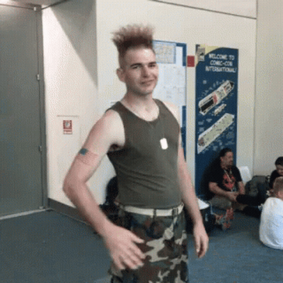 A Man In A Tank Top And Camo Pants Is Standing In Front Of A Welcome To Comic Con International Sign GIF
