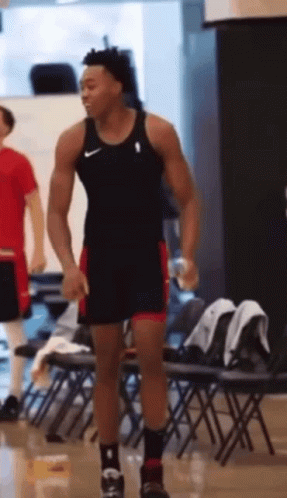 A Man In A Tank Top And Shorts Is Standing In Front Of A Row Of Folding Chairs . GIF