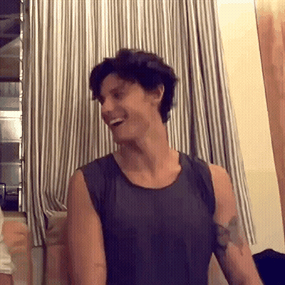 A Man In A Tank Top Is Laughing In Front Of A Striped Curtain In A Room . GIF