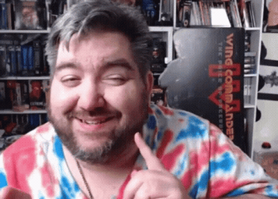 A Man In A Tie Dye Shirt Is Smiling In Front Of A Wing Commander Sign GIF