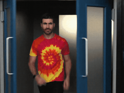 A Man In A Tie Dye Shirt Is Walking Through A Doorway GIF