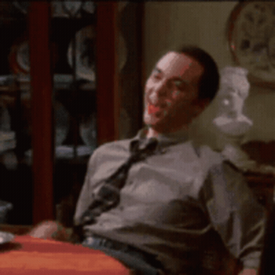 A Man In A Tie Is Sitting At A Table With A Clock In The Background GIF