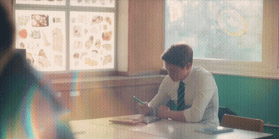 A Man In A Tie Sits At A Desk In A Classroom Looking At His Phone GIF