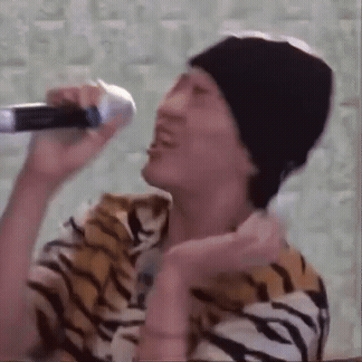 A Man In A Tiger Print Shirt Is Singing Into A Microphone While Wearing A Beanie . GIF
