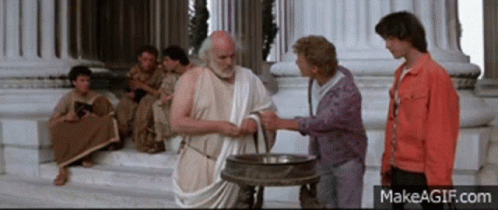 A Man In A Toga Shakes Hands With A Woman In A Red Jacket GIF