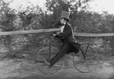 A Man In A Top Hat And Bow Tie Is Riding A Bicycle . GIF