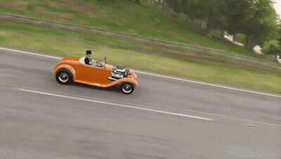 A Man In A Top Hat Drives An Orange Car On A Road GIF