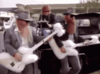 A Man In A Top Hat Is Playing A Guitar In Front Of A Car . GIF