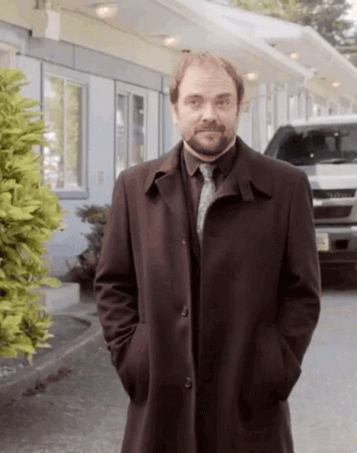A Man In A Trench Coat And Tie Is Standing In Front Of A Motel . GIF
