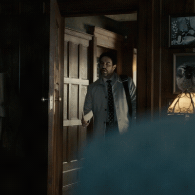 A Man In A Trench Coat And Tie Stands In A Hallway GIF