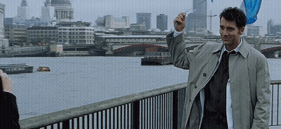 A Man In A Trench Coat And Tie Stands On A Bridge Overlooking A River GIF