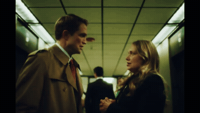A Man In A Trench Coat And Tie Talks To A Woman GIF