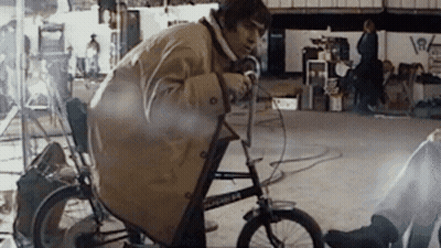 A Man In A Trench Coat Is Riding A Bicycle With The Number 64 On The Front Wheel GIF
