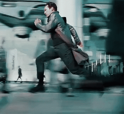 A Man In A Trench Coat Is Running Down A City Street GIF