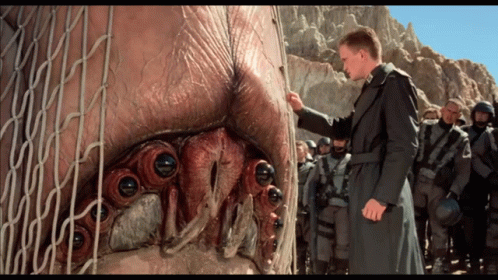 A Man In A Trench Coat Is Standing In Front Of A Large Monster GIF