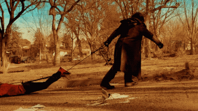 A Man In A Trench Coat Pulls A Person On A Leash GIF