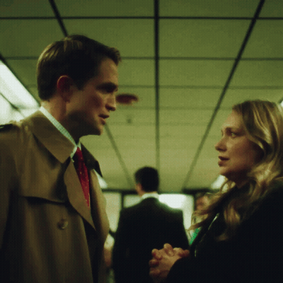 A Man In A Trench Coat Talks To A Woman In A Black Coat GIF