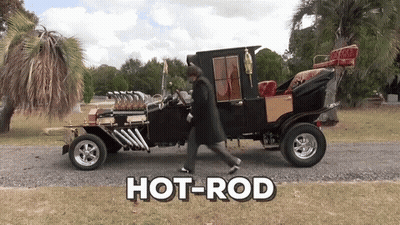 A Man In A Trench Coat Walks By A Hot Rod GIF