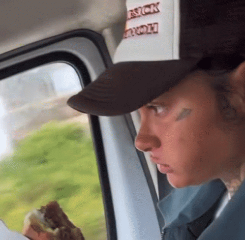 A Man In A Trucker Hat Is Eating A Hamburger While Looking Out The Window . GIF